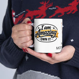 Person holding a mug with 'I am Amazing, Own It' text, wearing a dark sweater with red deer designs.