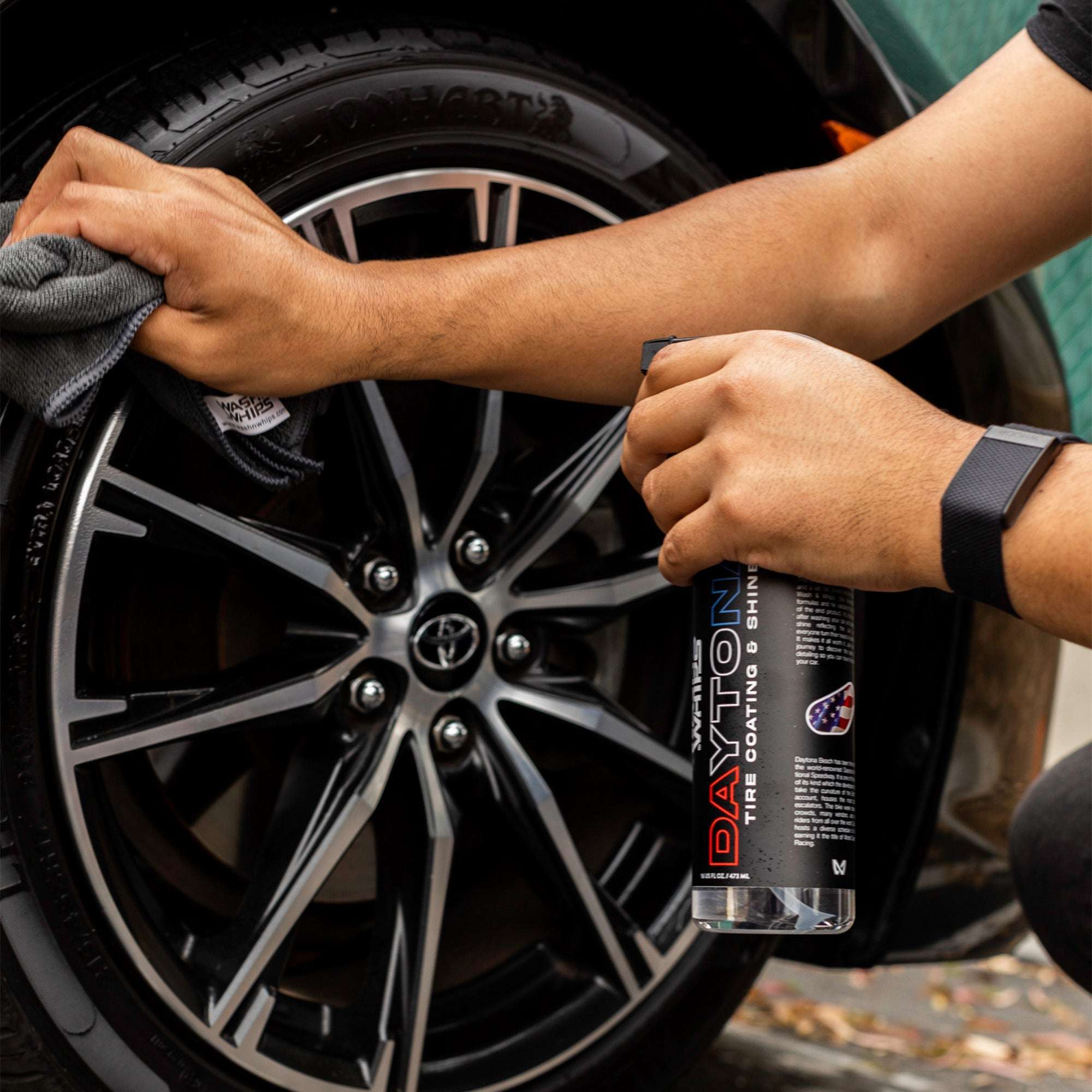 Person cleaning a car wheel with a cloth and holding a bottle of car care product.