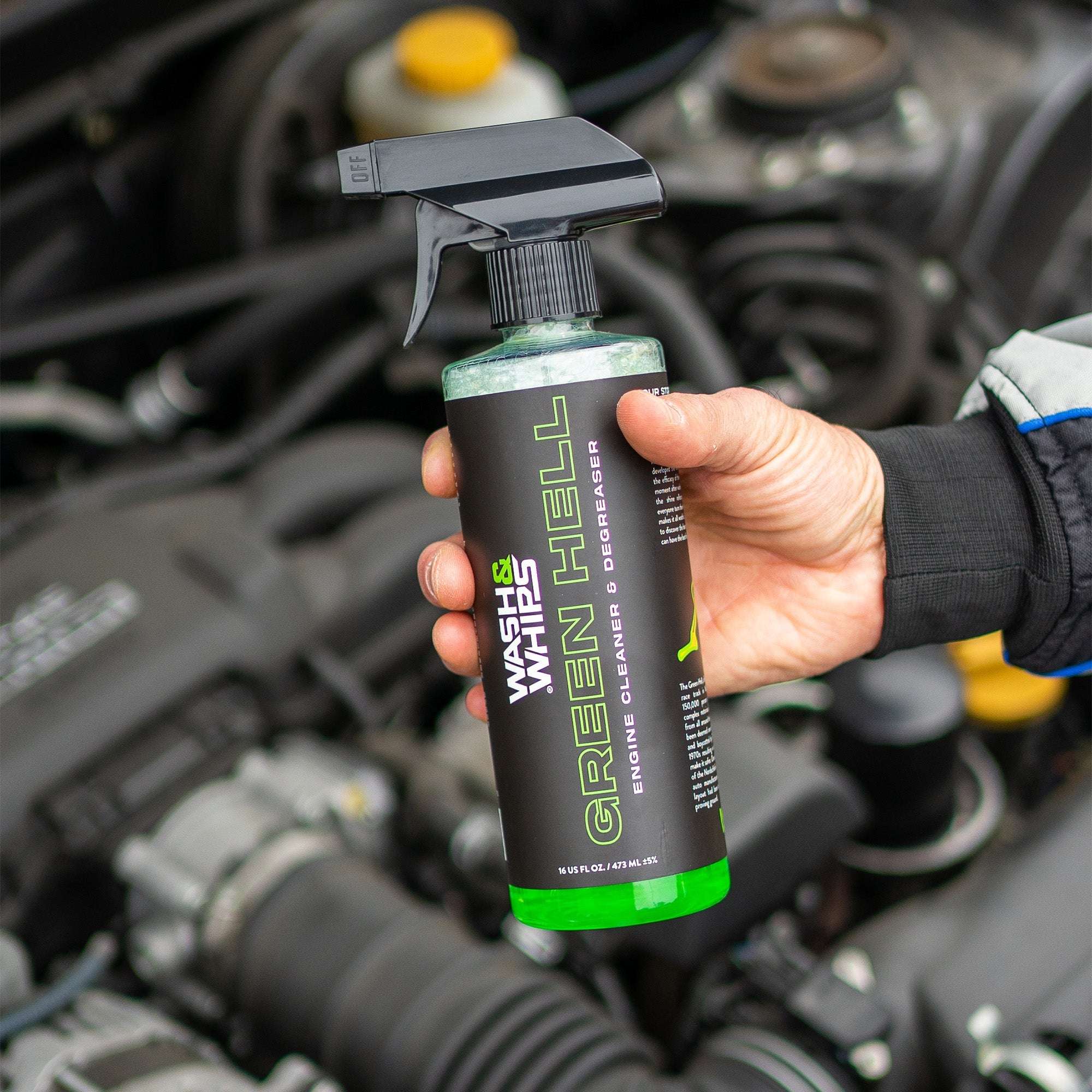 Hand holding a bottle of 'Green Hell' engine cleaner in front of a car engine.