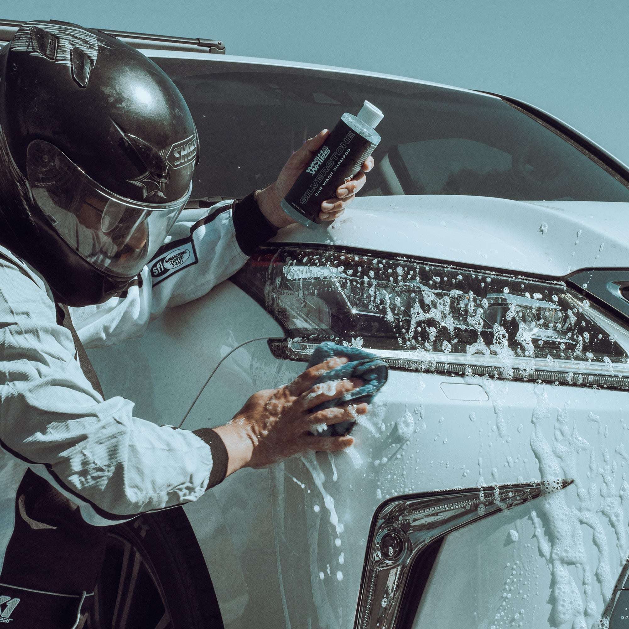 Person cleaning a car with a sponge and spray bottle, wearing a helmet and racing suit.