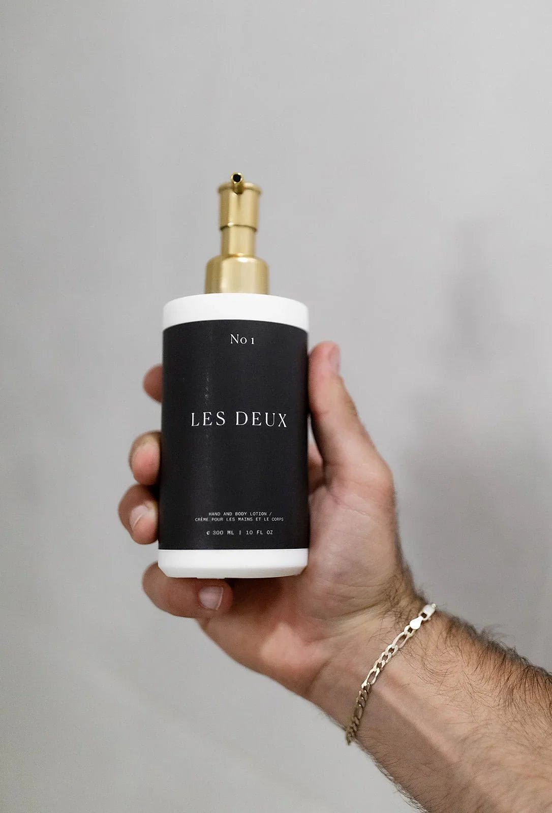 Hand holding a 'Les Deux' bottle with a gold pump against a gray background