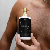 Hand holding a black and gold bottle labeled 'Les Deux' against a neutral background